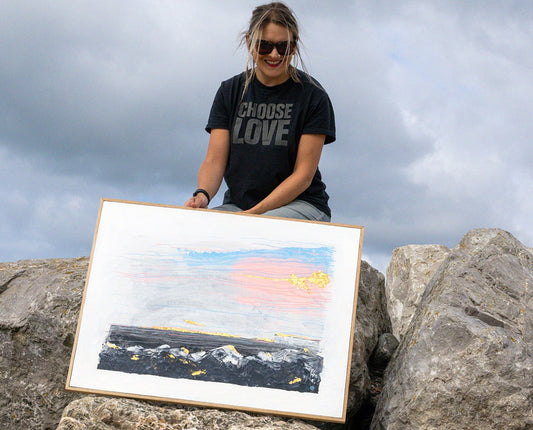 Jo Conlon sat on rocks on the beach, smiling with her painting rain or shine 